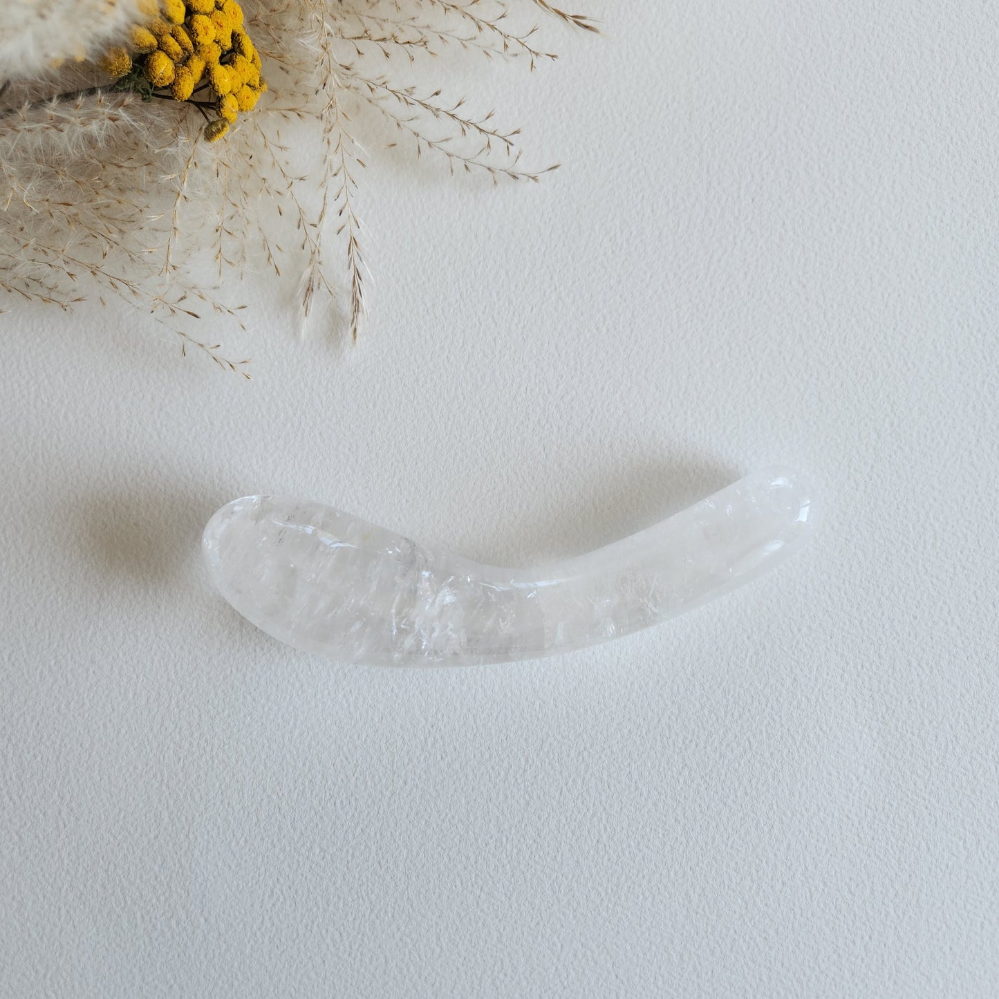 Smooth, curved, dual-sized clear quartz crystal wand with one bulbous and one tapered end, lying on a light surface with dried grasses and yellow flowers.