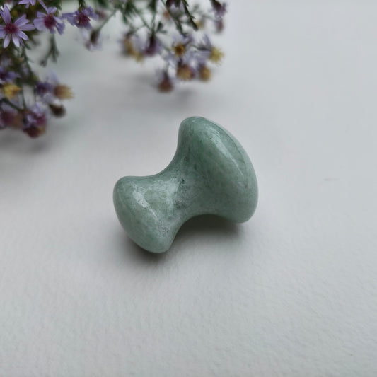 A small polished light green aventurine mushroom massager with a rounded, organic shape, resting on a white textured surface with purple wildflowers in the upper left corner.