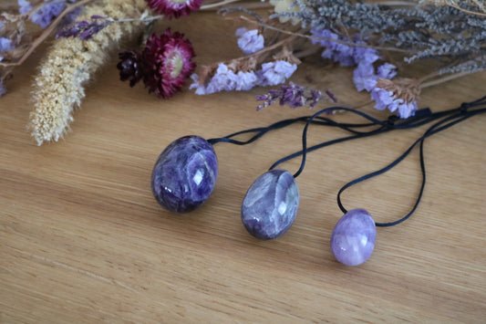 A set of three polished Amethyst yoni eggs in varying shades of purple, with black cords, lying on a wooden surface with dried flowers.
