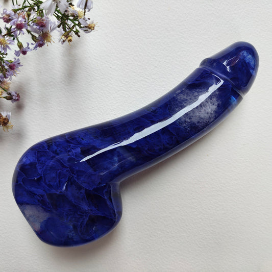 A glossy melting blue stone dildo with natural marbling, displayed diagonally on a textured off-white background with small wildflowers in the top-left corner.