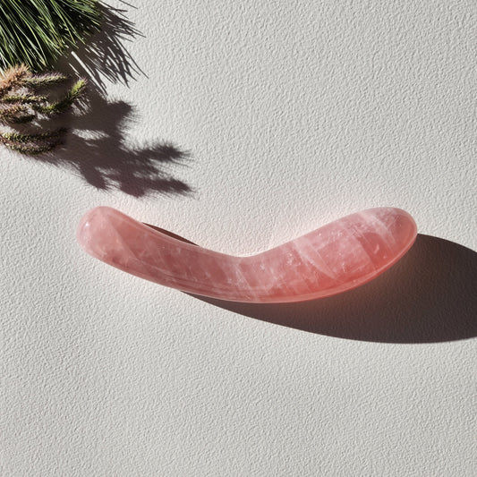 Large, curved, dual-sized rose quartz crystal wand with one thick and one thin end, lying on a light surface with pine needles and shadows.