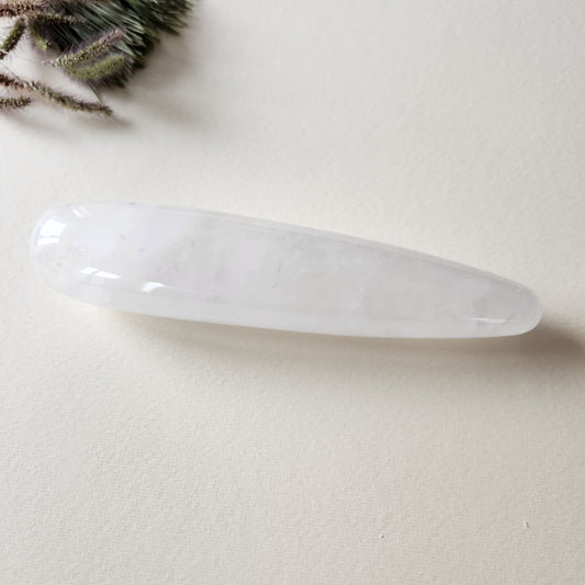 Polished, clear quartz yoni wand (large, straight) with subtle internal veining, on a cream-colored background. Natural light highlights the smooth, reflective surface of the crystal.