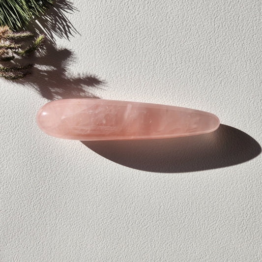 Polished Rose Quartz Yoni Wand (large, straight) with a soft pink hue and natural internal striations. The crystal rests on a light-colored background, with a sprig of pine needles casting a shadow. Perfect for sexual stimulation