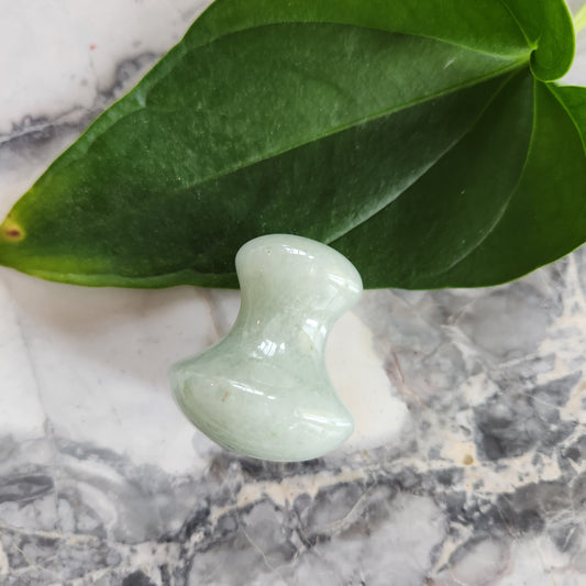 Small, polished green aventurine crystal massager with a dumbbell or mushroom shape, placed on a marble surface next to a large green leaf.