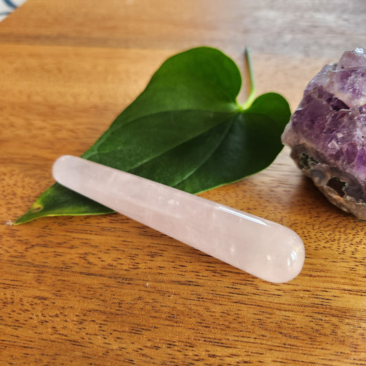 Small, smooth pink rose quartz crystal wand with a rounded end, placed on a wooden surface next to a green leaf and a rough amethyst geode.