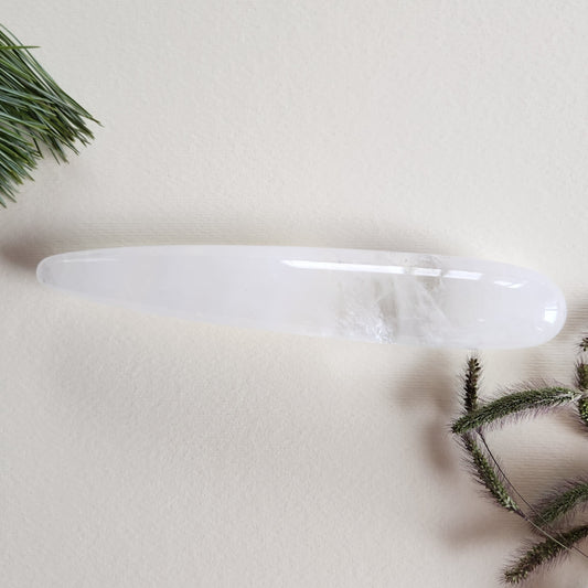 Polished clear quartz crystal yoni wand with natural internal variations and a smooth, tapered shape to reach the G-Spot, displayed on a light beige surface with pine needles and dried foliage.