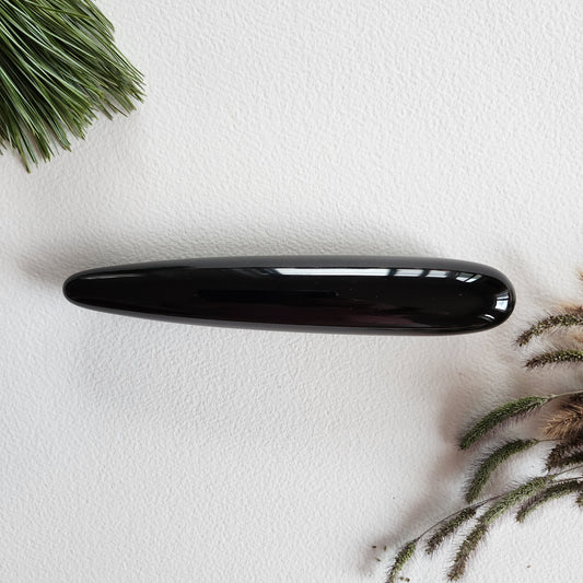 Polished black obsidian crystal yoni wand with a smooth, tapered shape for sex anal, shown on a white surface with pine needles and dried foliage