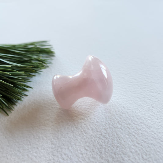 Small, polished rose quartz crystal massager with a dumbbell or mushroom shape, placed on a white surface next to a pine sprig.