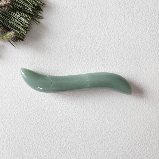 Small, S-shaped green aventurine crystal wand with a smooth, polished finish, lying on a white textured surface with natural pine needles.