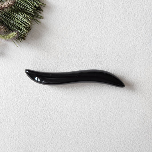 Small, S-shaped black obsidian crystal wand with a smooth, polished finish, lying on a white textured surface with natural pine needles.