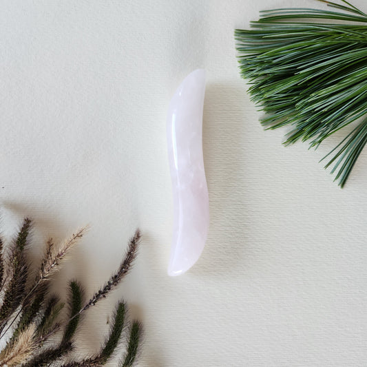 Small, S-shaped rose quartz crystal wand with a smooth, polished finish, lying on a white textured surface with natural pine needles and dried foliage.