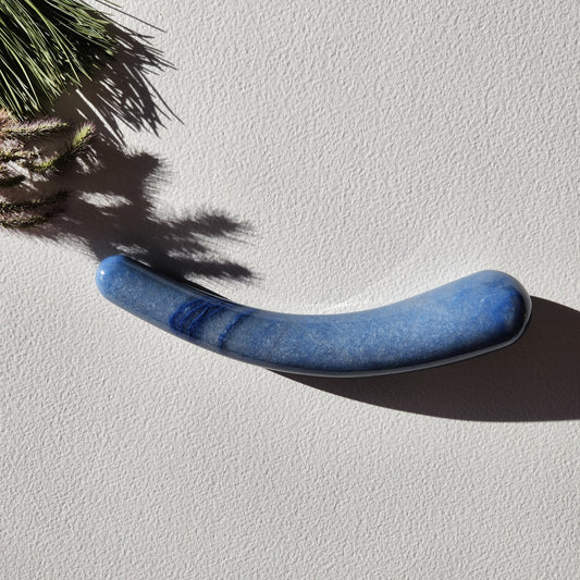 Smooth, crescent-shaped blue aventurine crystal wand with a uniform thickness, lying on a light surface with pine needles and shadows from a plant.