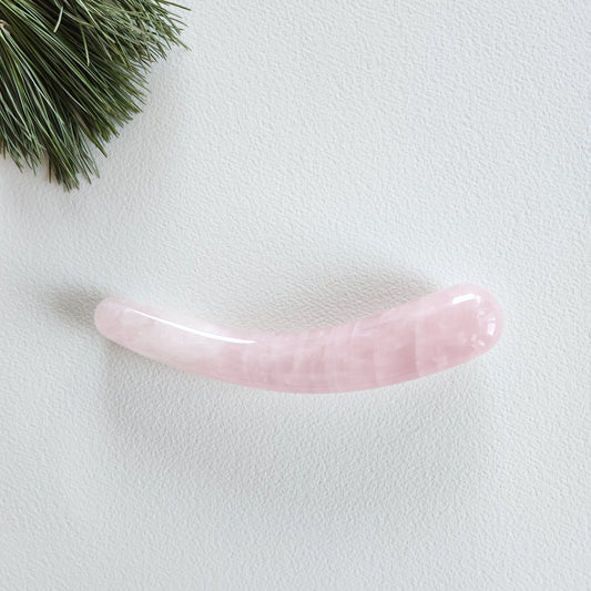 Smooth, crescent-shaped rose quartz crystal wand with a uniform thickness, lying on a white surface with pine needles and shadows from a plant.