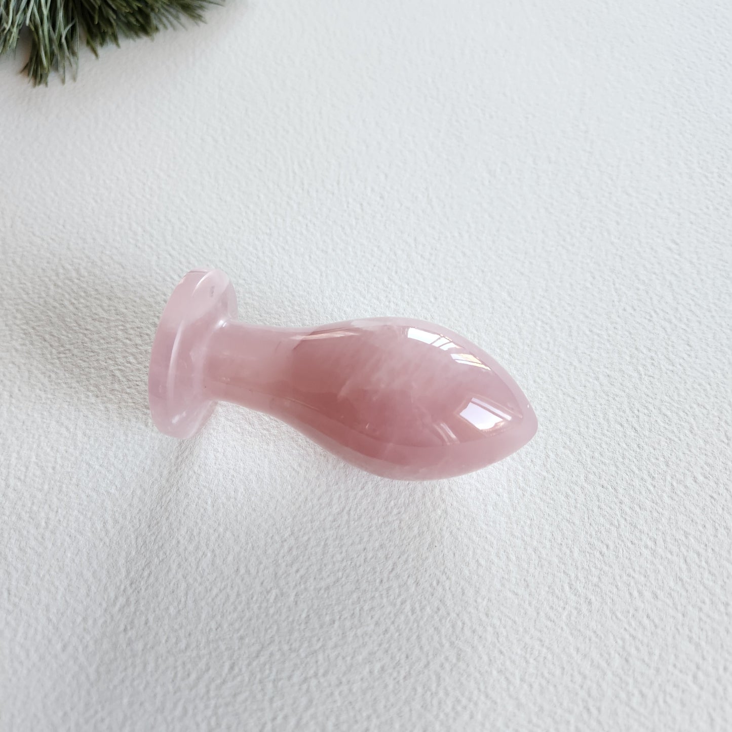 A hand-polished, curved rose quartz Butt-Plug lying on a light-colored surface with a green pine branch in the background.