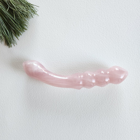 Smooth, double-ended rose quartz crystal wand with a gently curved end and a beaded or segmented end, lying on a white surface with pine needles.