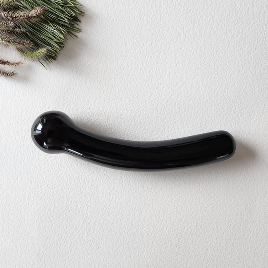 A hand-polished, curved black obsidian crystal wand with round tip lying on a light-colored surface with a green pine branch in the corner.