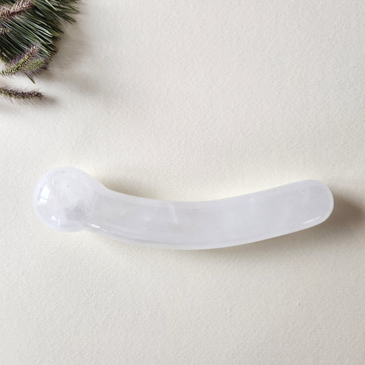 Curved clear quartz crystal wand with a smooth, polished finish, designed for G-spot stimulation, lying on a light surface with pine needles and dried foliage.