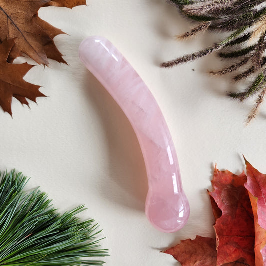 Curved rose quartz crystal wand with a smooth, polished finish, designed for G-spot stimulation, lying on a light surface surrounded by autumn leaves and pine needles.
