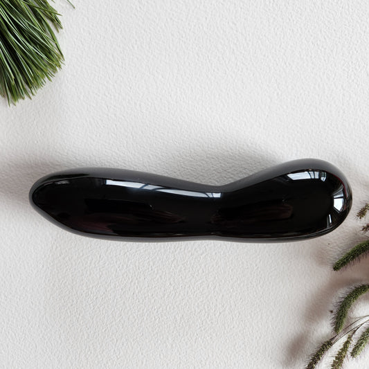 Large, polished black obsidian crystal wand with a unique contoured, spoon-like shape, lying on a white surface with pine needles and dried foliage.