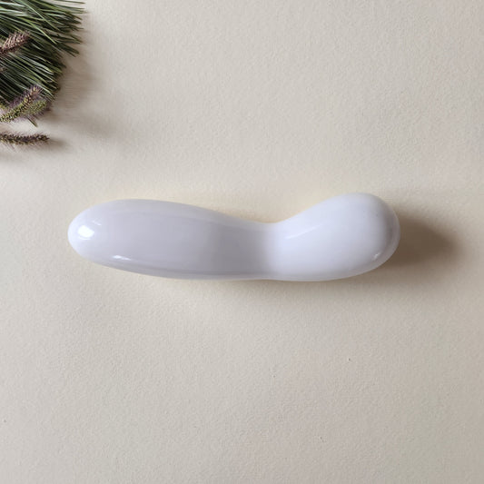 Large, polished white jade crystal wand with a unique contoured, spoon-like shape, lying on a light surface with pine needles and dried foliage.