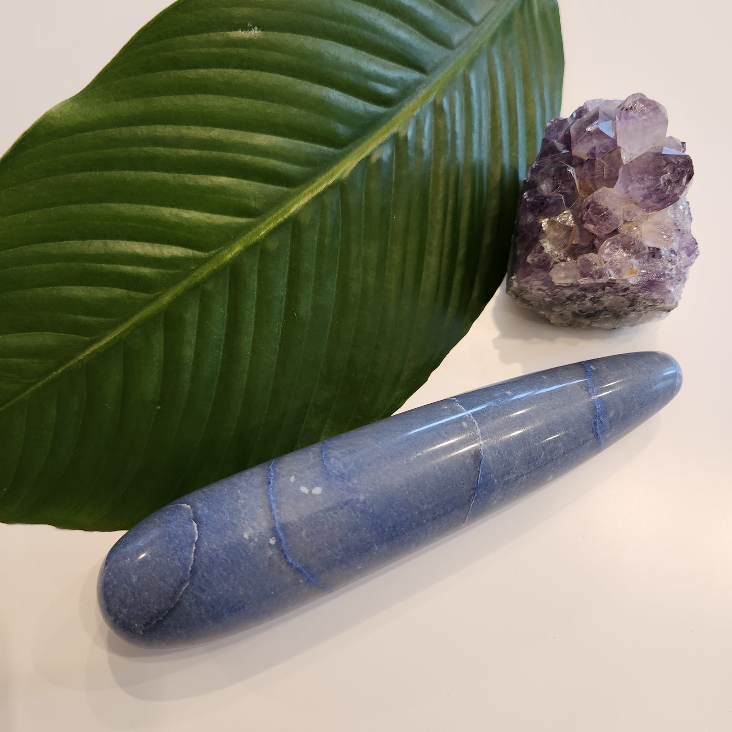 Thick, polished blue aventurine crystal wand with a large diameter perfect for anal pleasure, lying on a white surface next to a large green leaf and a rough amethyst geode.