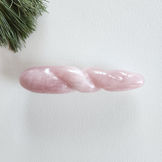 Polished rose quartz yoni wand with a twisted, spiral body and a pointed tip, lying on a white surface with pine needles.