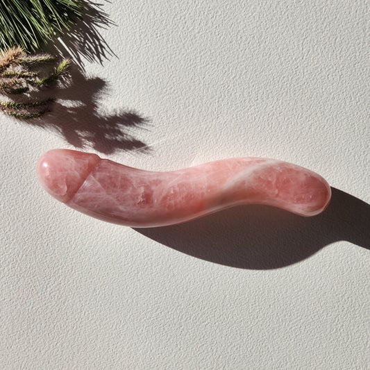 Polished rose quartz wand with a phallic shape and a distinct S-curve, lying on a white surface with pine needles and a strong shadow.