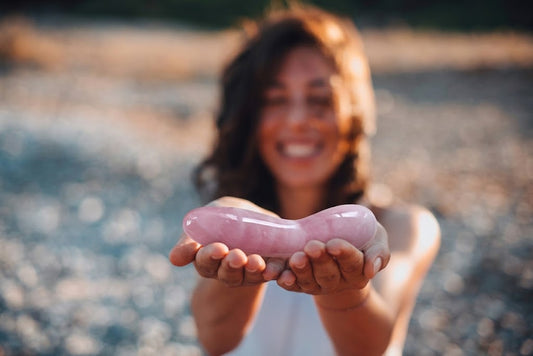 A woman holding the Rose Quartz Large Curved yoni wand in her open palms at golden hour on a pebbled beach