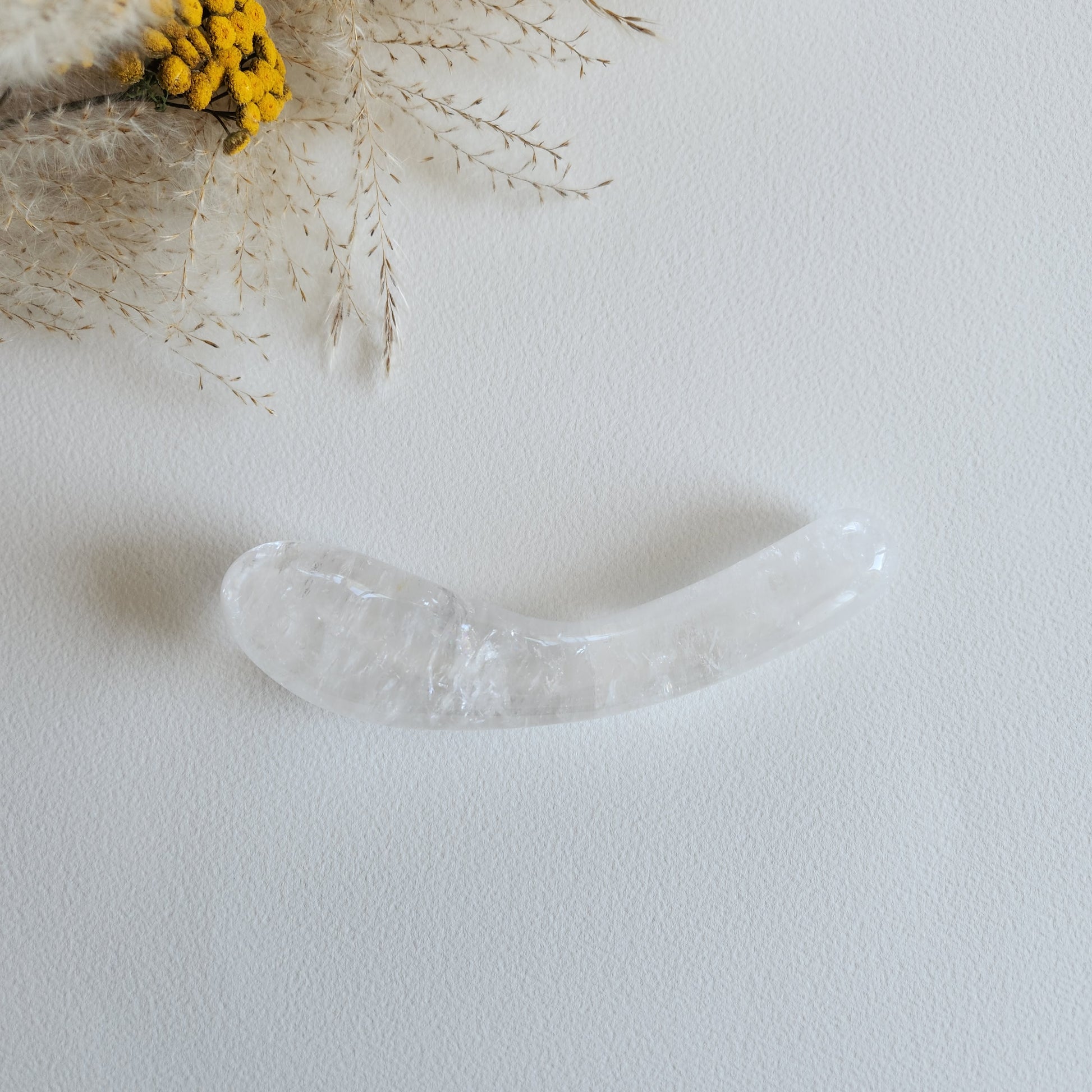 Smooth, curved, dual-sized clear quartz crystal wand with one bulbous and one tapered end, lying on a light surface with dried grasses and yellow flowers.