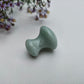 A small polished light green aventurine mushroom massager with a rounded, organic shape, resting on a white textured surface with purple wildflowers in the upper left corner.