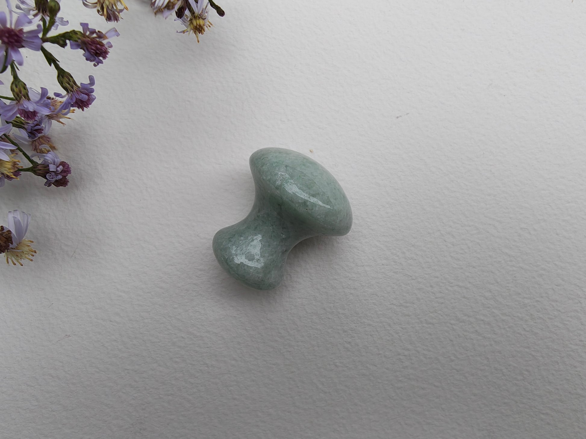A small mushroom-shaped massager made of polished light green aventurine stone, shown on a textured white background with purple wildflowers in the top-left corner.