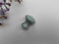 A small mushroom-shaped massager made of polished light green aventurine stone, shown on a textured white background with purple wildflowers in the top-left corner.