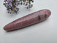 A polished rhodonite stone cone with natural dark veining, photographed on a light surface with small purple flowers in the upper background.