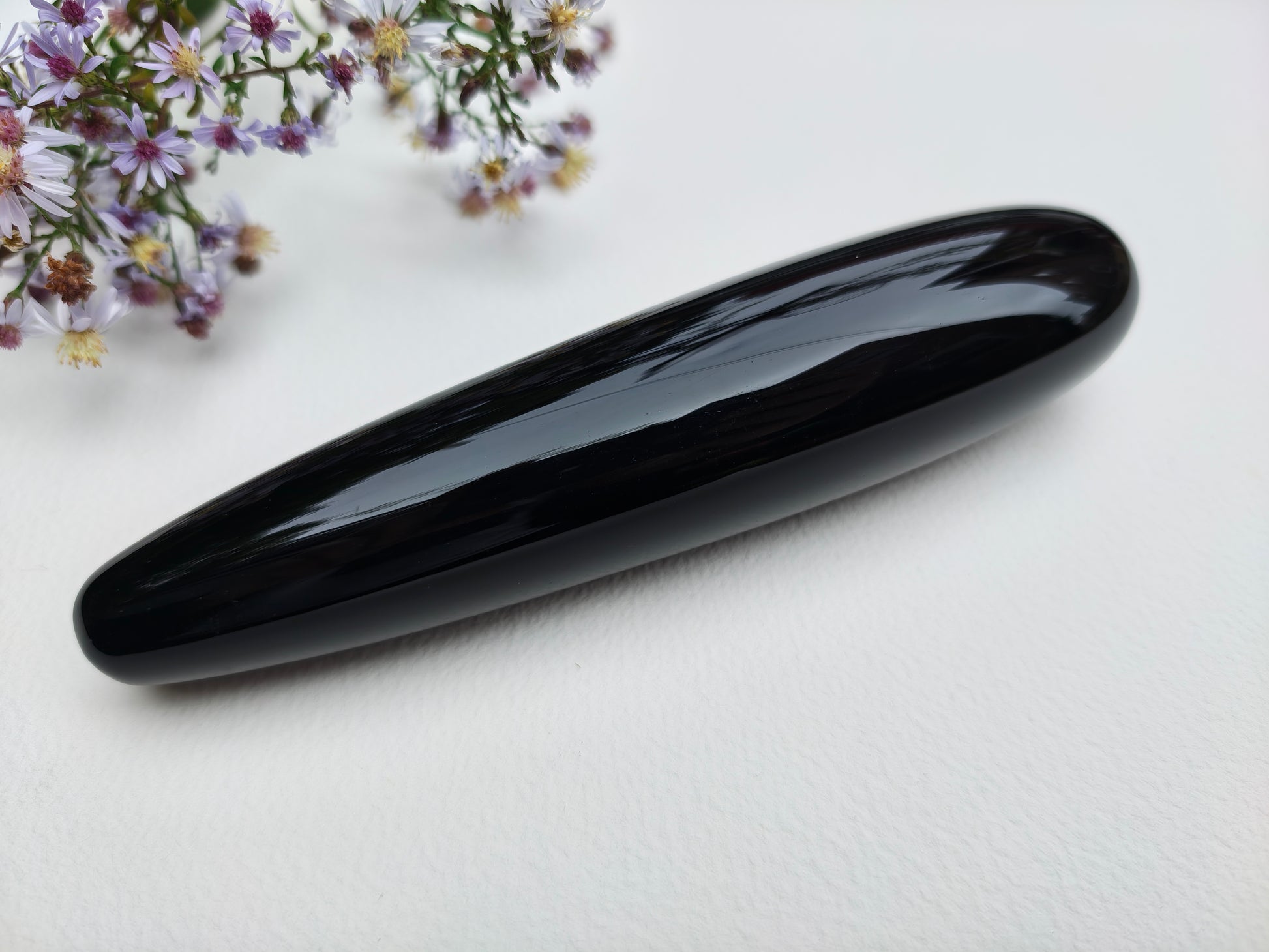 A polished black obsidian stone tool with a smooth, glossy finish, resting on a white textured surface with small purple flowers in the top-left corner.