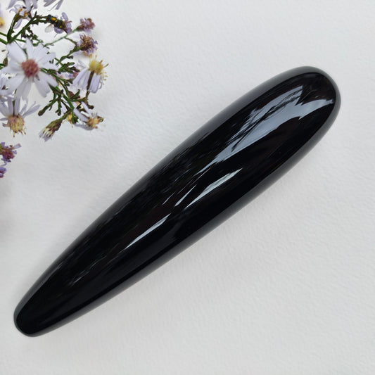 A glossy black obsidian, elongated oval polished stone, displayed diagonally on a textured white surface next to purple wildflowers.