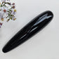 A glossy black obsidian, elongated oval polished stone, displayed diagonally on a textured white surface next to purple wildflowers.