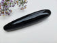 A polished black obsidian stone tool with a smooth, glossy finish, resting on a white textured surface with small purple flowers in the top-left corner.