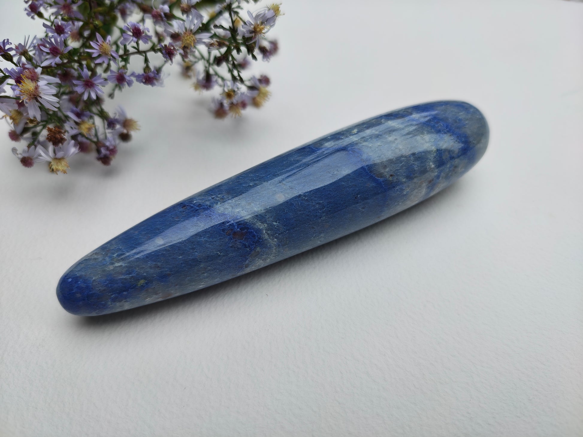 A polished blue lapis lazuli crystal point with natural white and golden matrix inclusions, resting on a white textured surface next to small purple wildflowers.