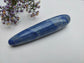 A polished blue lapis lazuli crystal point with natural white and golden matrix inclusions, resting on a white textured surface next to small purple wildflowers.