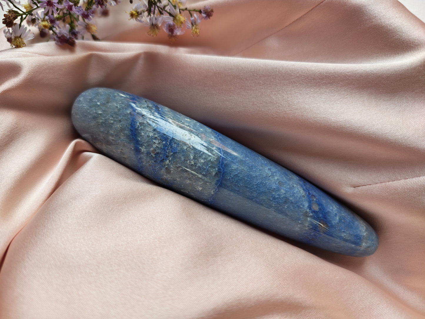A polished blue stone cylinder, blue aventurine crystal wand, resting on draped blush pink satin fabric with small wildflowers in the corner.