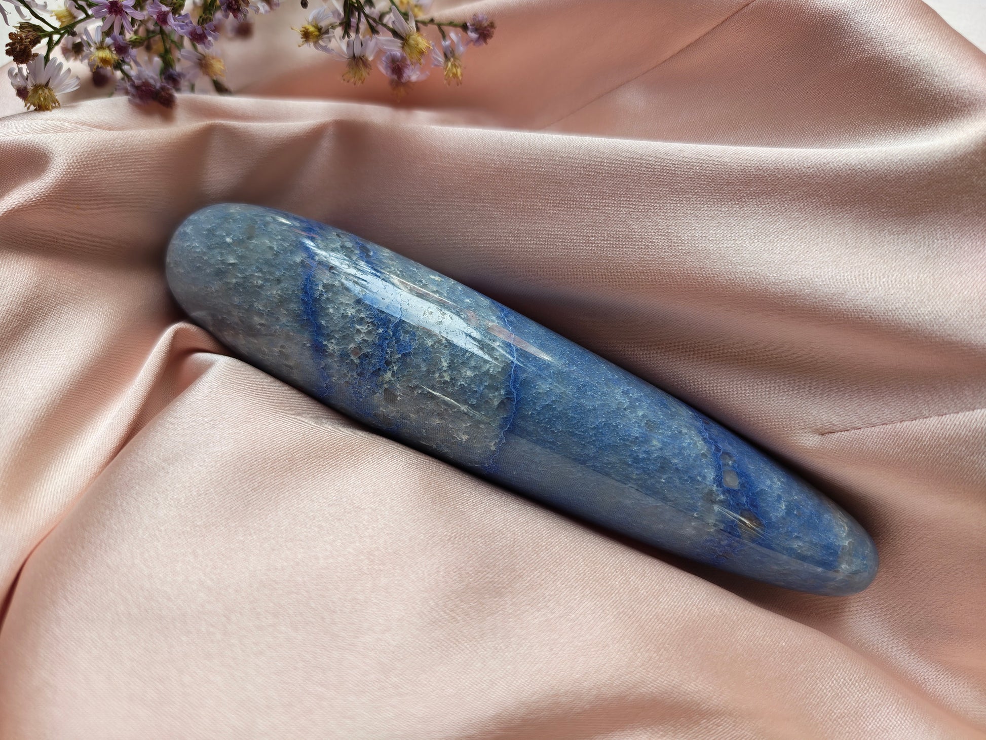 A polished blue stone cylinder, blue aventurine crystal wand, resting on draped blush pink satin fabric with small wildflowers in the corner.