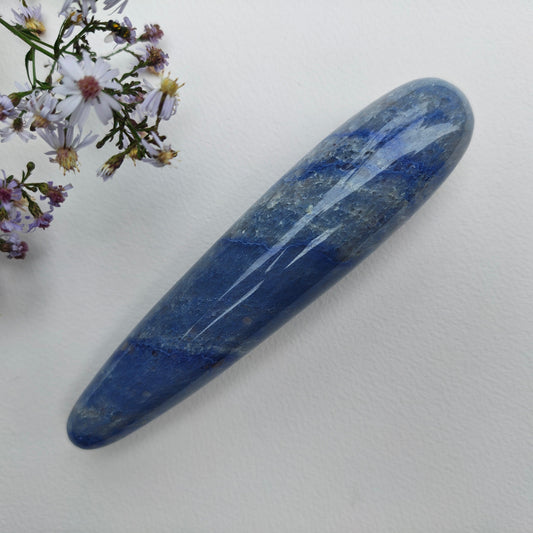 A polished blue lapis lazuli crystal point with natural white and gold veining, displayed diagonally on a textured white surface next to white and purple wildflowers.