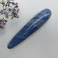 A polished blue lapis lazuli crystal point with natural white and gold veining, displayed diagonally on a textured white surface next to white and purple wildflowers.