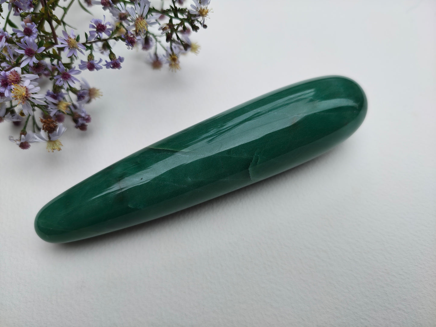 A polished green aventurine crystal yoni wand point resting on a white surface, with small purple flowers in the top-left corner.