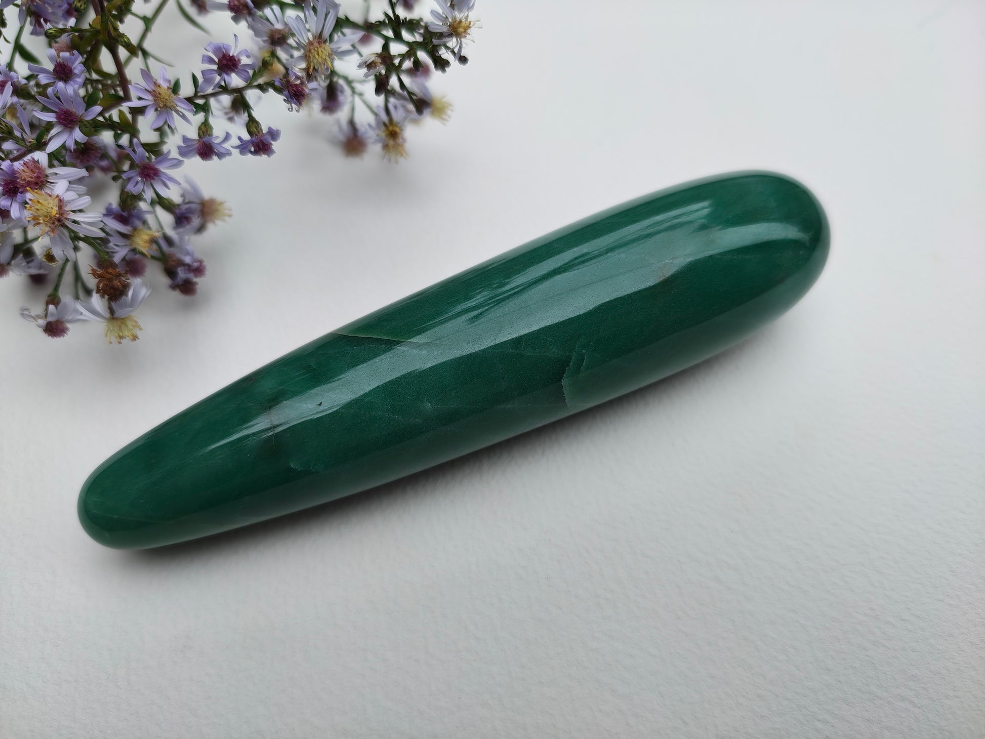 A polished green aventurine crystal yoni wand point resting on a white surface, with small purple flowers in the top-left corner.