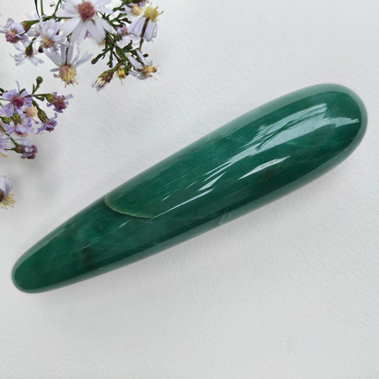 A polished green aventurine crystal wand, displayed diagonally on a white textured surface with small purple wildflowers in the top-left corner.