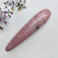 A polished pink stone wand, rhodonite, displayed diagonally on a white textured surface next to small purple and white flowers.