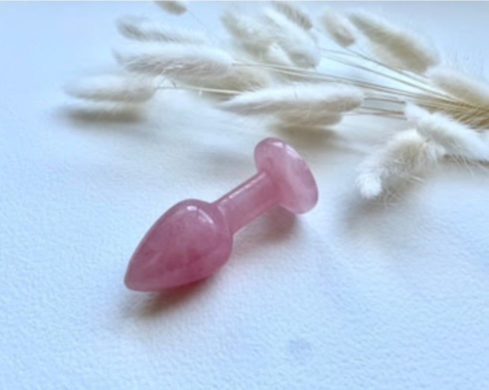 Small, polished rose quartz butt plug with a tapered tip and flat base, lying on a white surface with a bundle of dried, fluffy plant material.