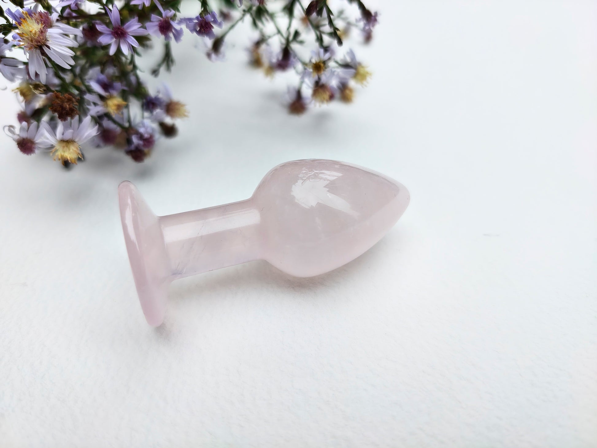 A pale pink rose quartz butt plug with a smooth, polished finish, resting on a white textured surface with dried purple flowers in the background.