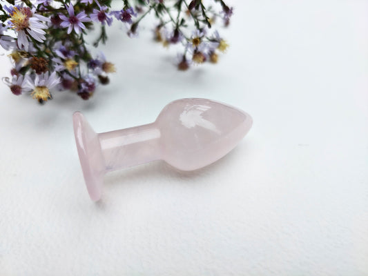 A pale pink rose quartz butt plug with a smooth, polished finish, resting on a white textured surface with dried purple flowers in the background.